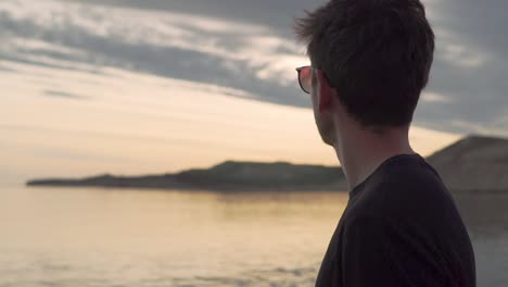 young man wearing sunglasses looking at the calm ocean during sunset