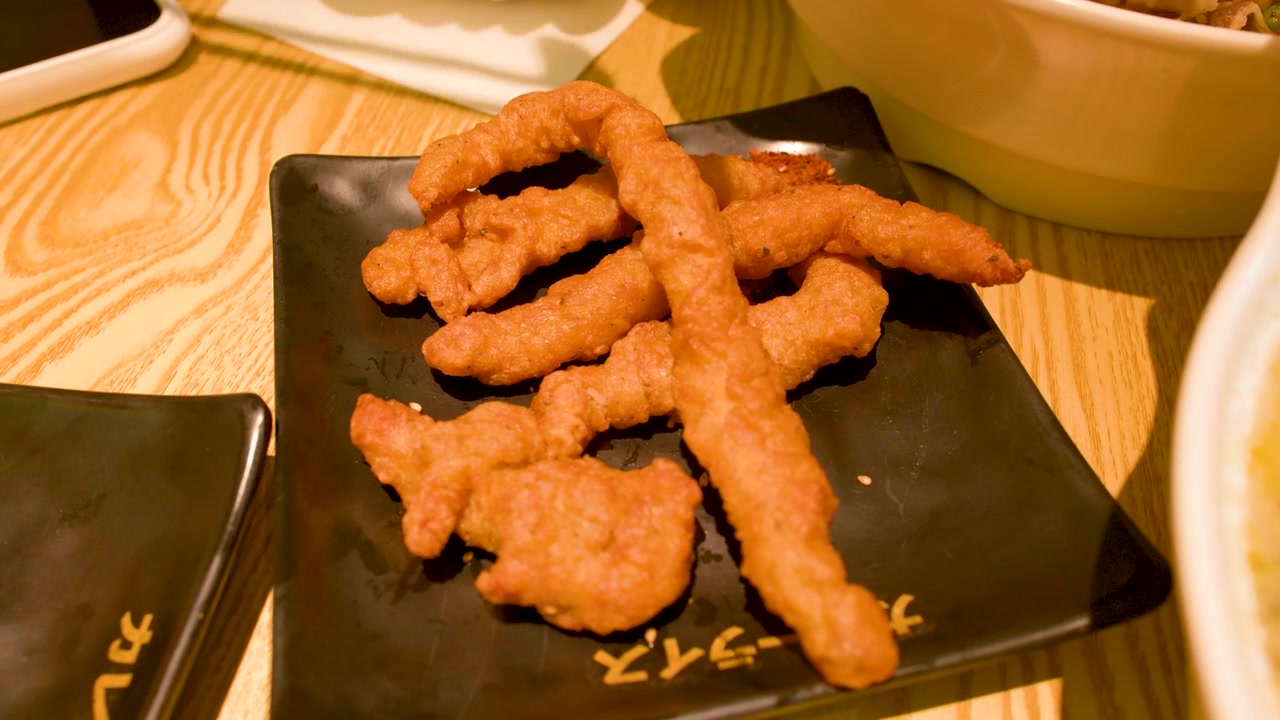 Plano detalhe de pauzinhos levantando frango frito dourado de um prato preto em um restaurante.