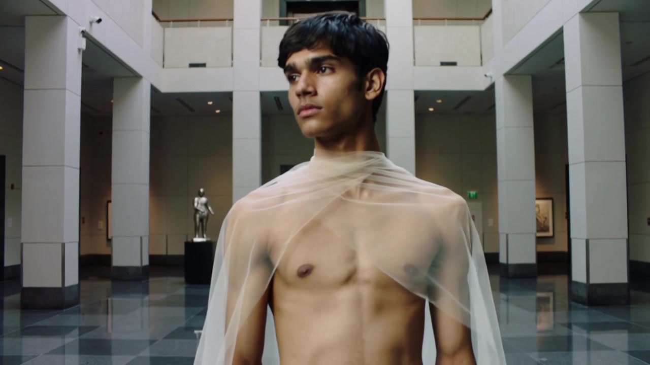 Modello a torso nudo che, guardando a sinistra, si gira e cammina lungo una galleria verso le sculture, con un mantello trasparente che strascica