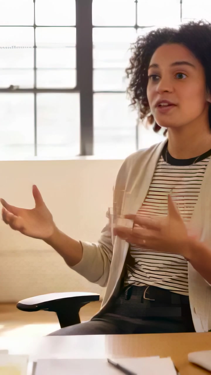 Vertical video: Starting to speak, woman in striped shirt explaining point at office desk with pen