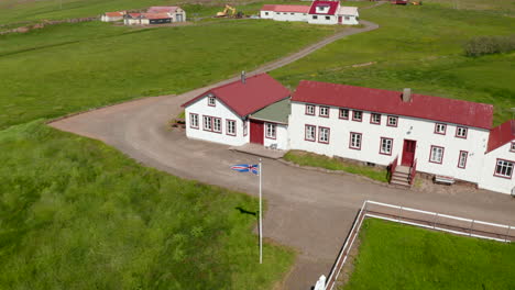 Drone-view-orbit-around-Iceland-flag-waving-out-of-typical-building-in-Iceland-highland.-The-flag-of-Iceland-fluttering-in-the-wind-in-a-small-settlement-in-Icelandic-countryside