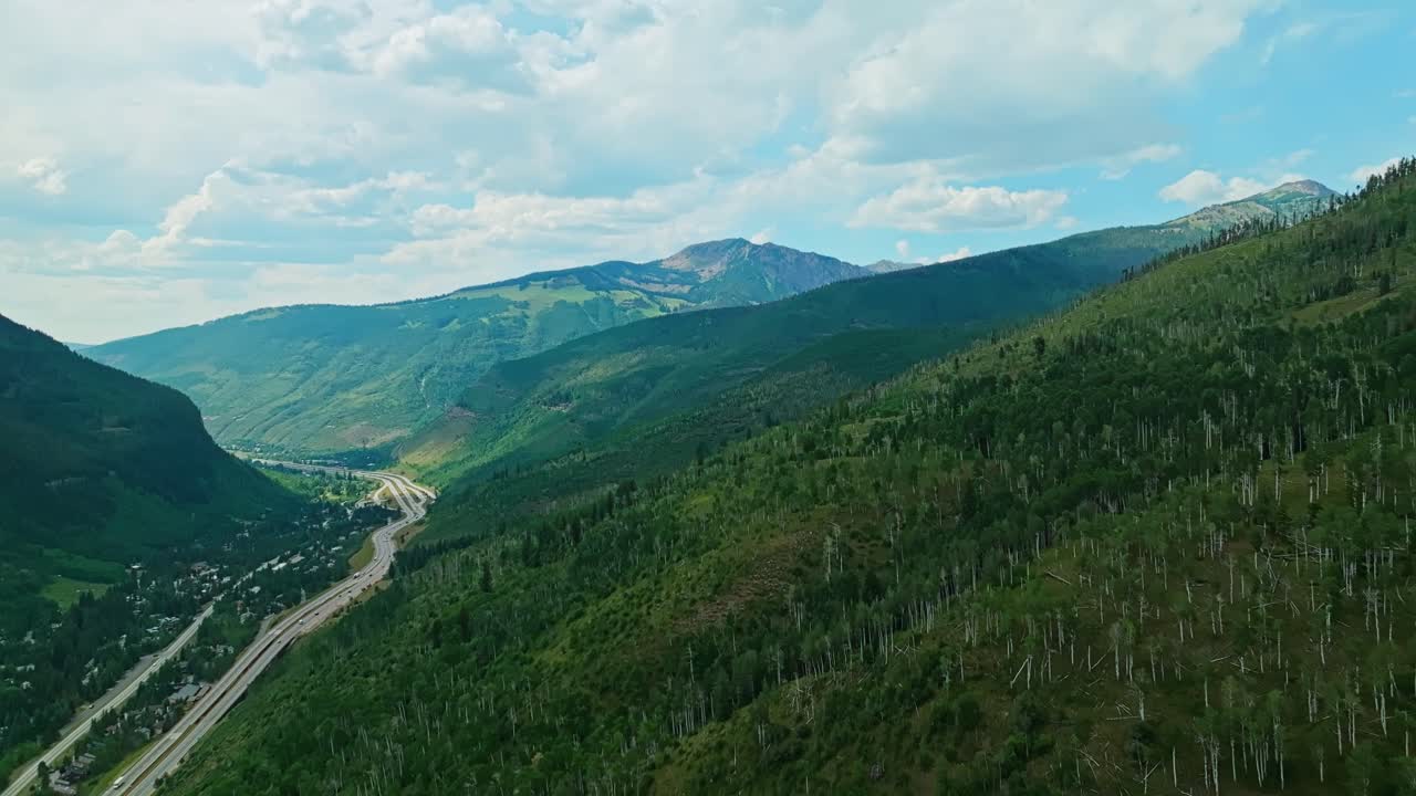 Aerial flyover of tree-lined Vail slope with gentle rolling and open alpine summer terrain, panoramic above highway