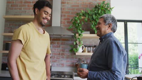 Cheerful-biracial-father-and-son-talking-while-toasting-coffee-mugs-in-kitchen-at-home