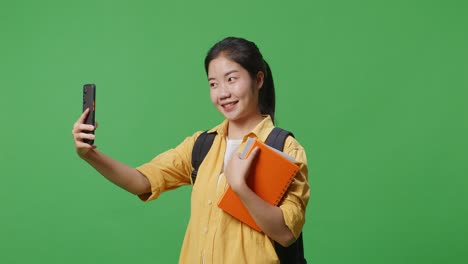 side view of asian woman student with a backpack and some books taking photo on smartphone while standing in the green screen background studio