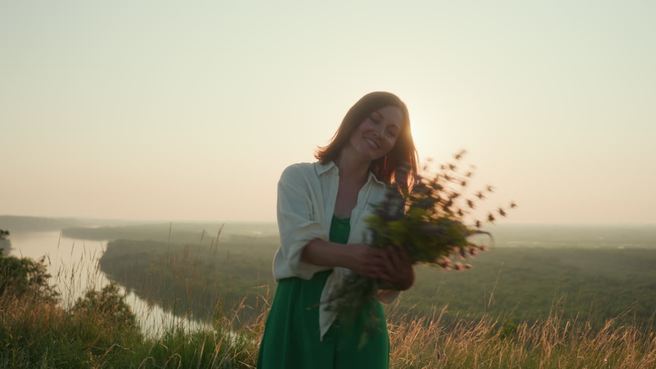 Sunset backlit young woman in green dress twirls joyfully on grassy river bank holding wildflower bouquet under warm golden light over river valley with shimmering sun glow and gentle breeze