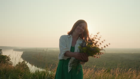 sunset backlit young woman in green dress twirls joyfully on grassy river bank holding wildflower bouquet under warm golden light over river valley with shimmering sun glow and gentle breeze