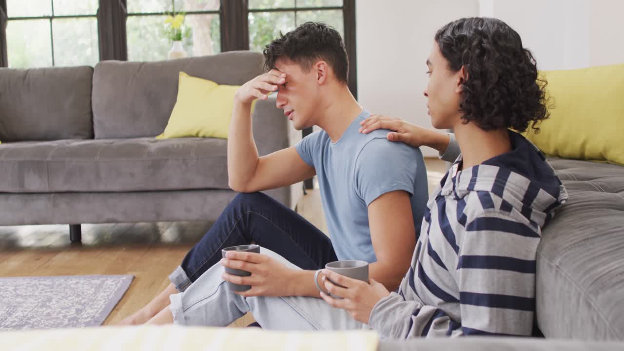 Worried diverse male couple sitting on floor and drinking coffee in living room