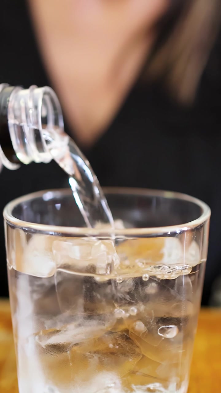 Una bottiglia trasparente versa acqua in un bicchiere pieno di cubetti di ghiaccio su un tavolo di legno, con una persona sfocata sullo sfondo e una luminosa illuminazione interna.
