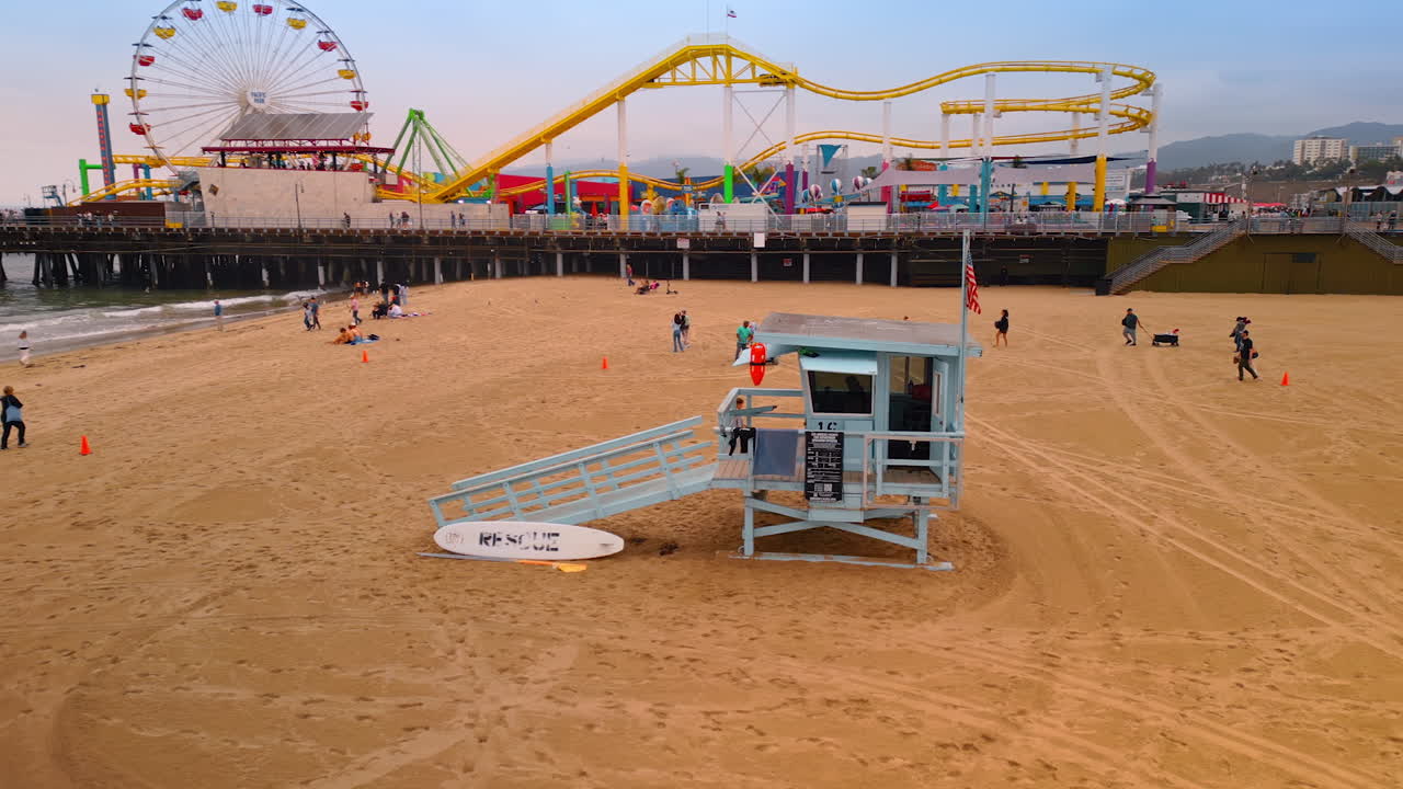 Los Angeles, USA, 29 August 2025: View on the lifeguard stand with an American flag on the sandy beach. Pacific Park on the Santa Monica Pier at backdrop. LA, California, USA from drone footage
