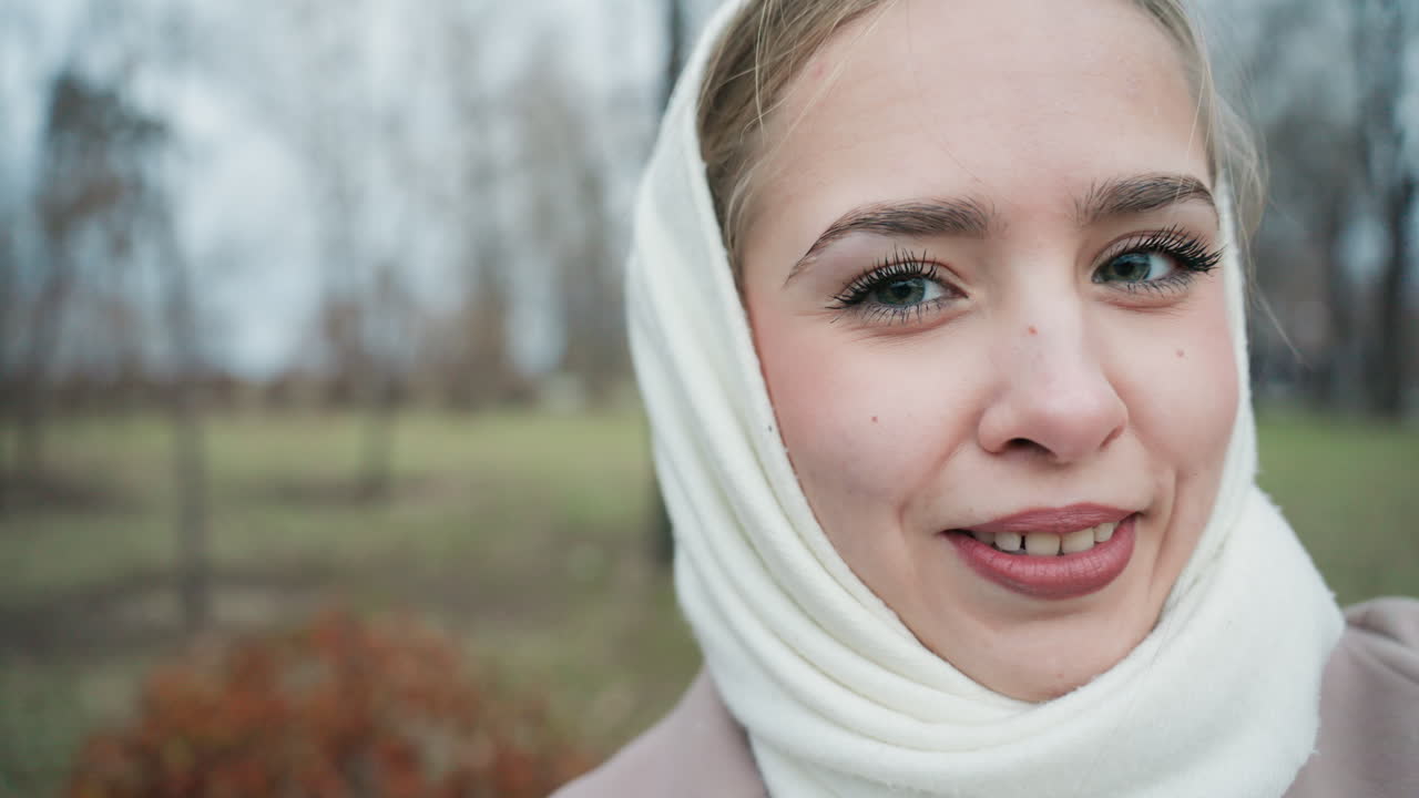 Young woman blowing kiss in park, white scarf frames face in intimate closeup with warm smile, soft bokeh trees behind, tender goodbye gesture and wistful eye contact, winter chill softened by cozy