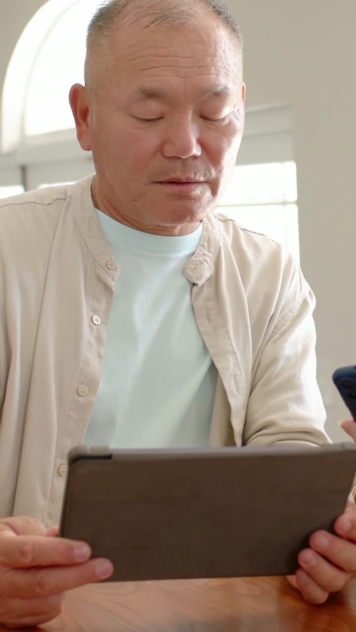縦型動画：自宅のテーブルでタブレットを立てかけ読書するベージュのシャツを着た高齢者、電話が鳴ってちらりと見る