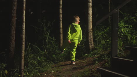 little boy in bright green tracksuit plays in evening forest pretending to be giant near wooden stairs. kid in comfortable clothes during family vacation in nature