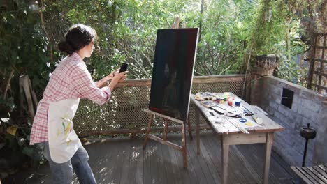 biracial female artist taking picture of her painting using smartphone in sunny garden, slow motion