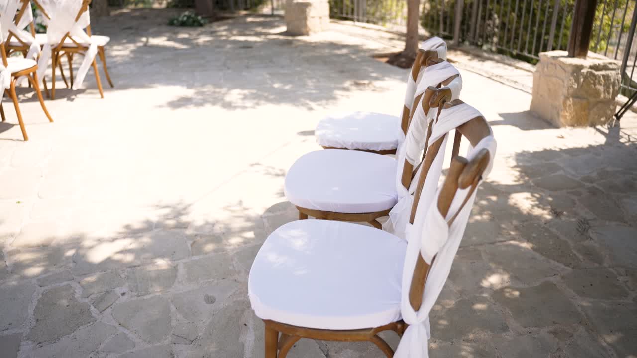Three wooden chairs are decorated with white fabric, set outside in a bright, sunny location. The scene features greenery and a relaxing atmosphere, perfect for gathering