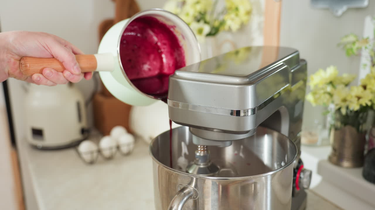 Hand holding mint green saucepan pours vibrant blended currant mixture into electric stand mixer bowl in bright kitchen setting with fresh eggs and flowers in soft background