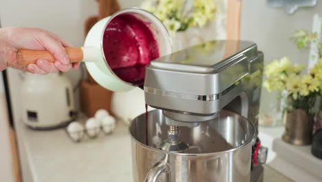 hand holding mint green saucepan pours vibrant blended currant mixture into electric stand mixer bowl in bright kitchen setting with fresh eggs and flowers in soft background