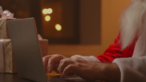 Hands-Santa-typing-on-wireless-keyboard-by-wooden-New-Year-decorated-table-Santa-Claus-is-working-with-a-laptop-looking-through-mail-and-answering-messages-to-children.-High-quality-4k-footage