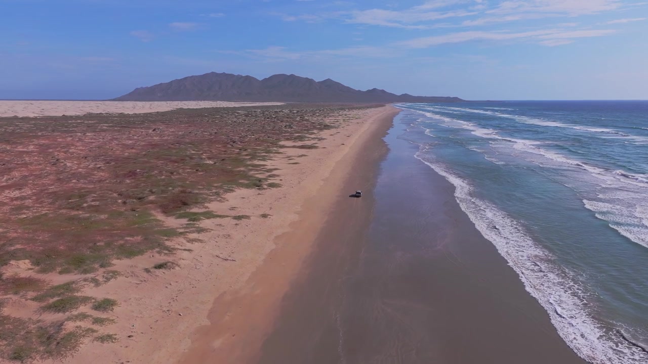 Vista de drone acompanhando um caminhão dirigindo ao longo da costa molhada de uma vasta praia deserta com dunas e montanhas ao fundo