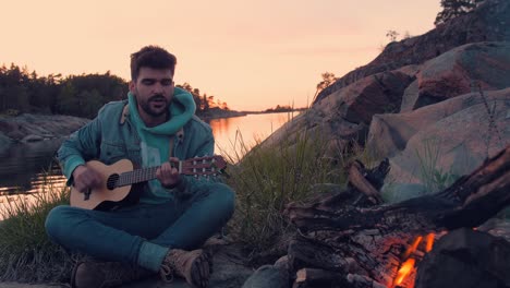 young man singing with acoustic guitar in front bonfire and sea on beautiful lonely island.