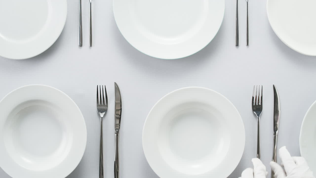 Minimalist table setting with white plate, fork, and knife arranged neatly, and a gloved hand adjusting cutlery.