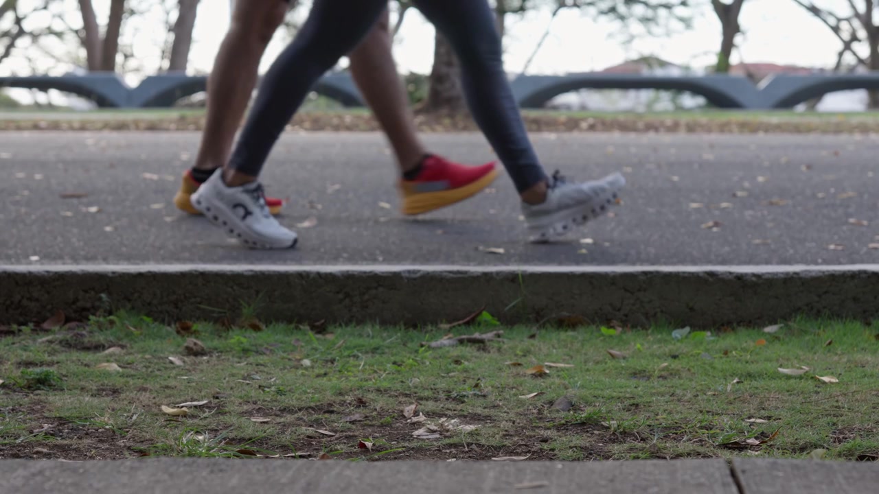 Pengambilan gambar statis dari permukaan tanah yang berfokus pada kaki dan sepatu atletik dua orang yang berlari lewat di jalan taman beraspal saat berolahraga di luar ruangan.