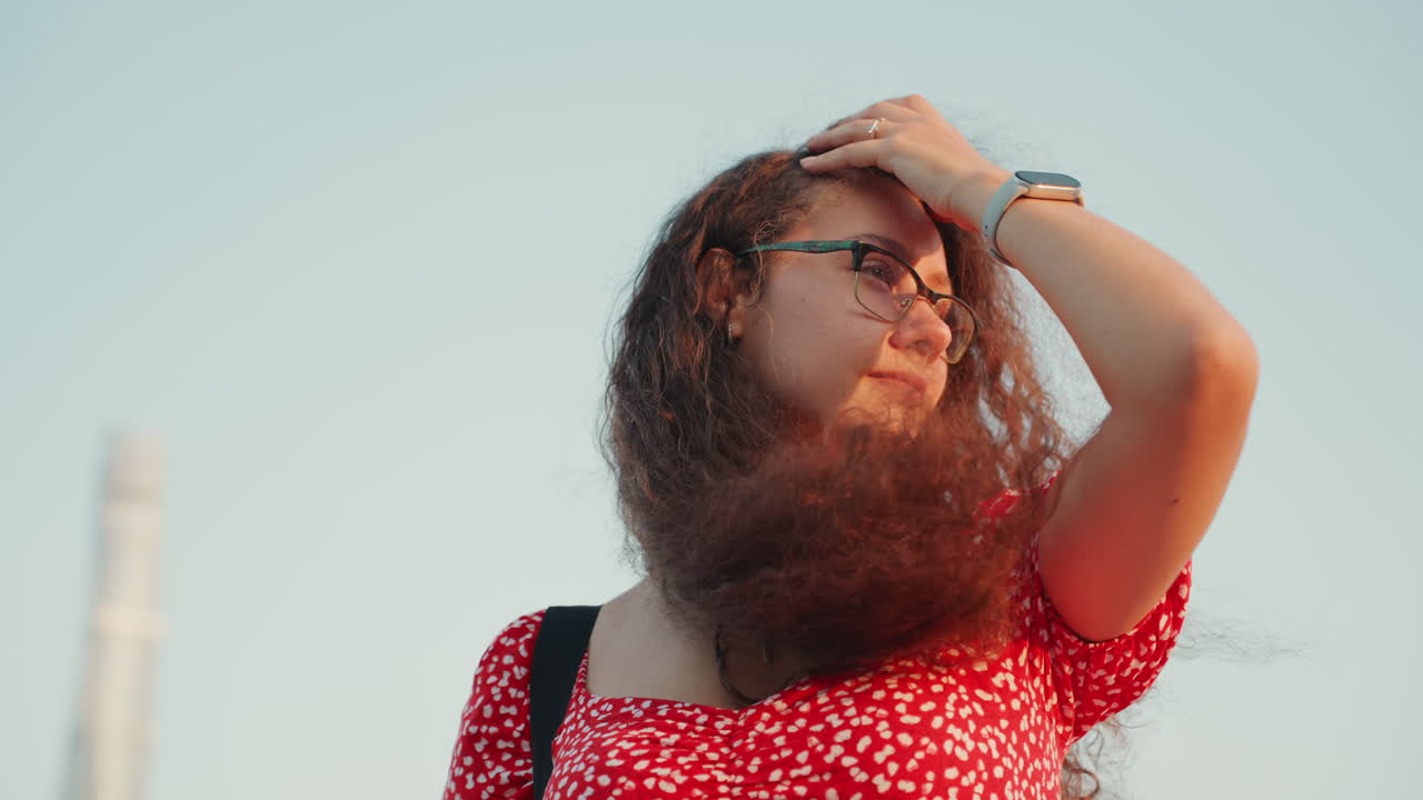 Joven mirando el horizonte al anochecer con cabello rizado barrido por el viento, usando gafas y un vestido floral rojo, una mano apartando el cabello en un gesto espontáneo, con una torre y un panorama de la ciudad detrás de ella.