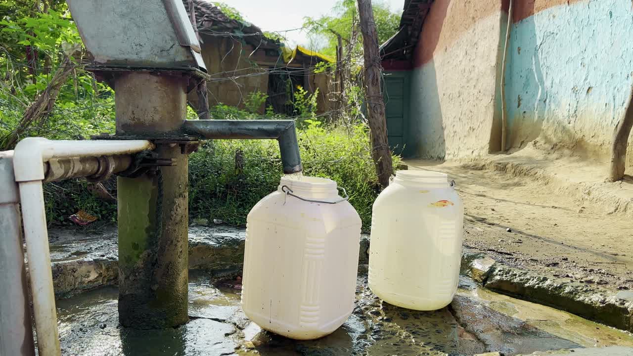 A person pumps water from a hand pump (off-frame) to fill two white cans in a rustic Indian village, symbolizing everyday life, community resilience, and access to essential natural resources