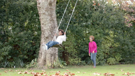 Happy african american mother pushing her son on tree swing in garden, in slow motion