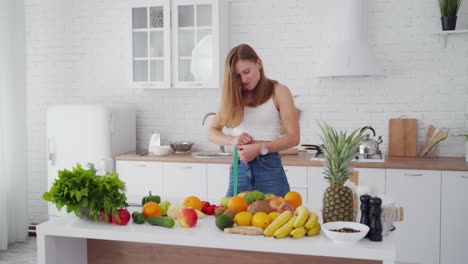 slim woman measures her waist in the kitchen. happy girl in jeans standing near the table full of fresh fruit and vegetables at home. young lady loses weight.