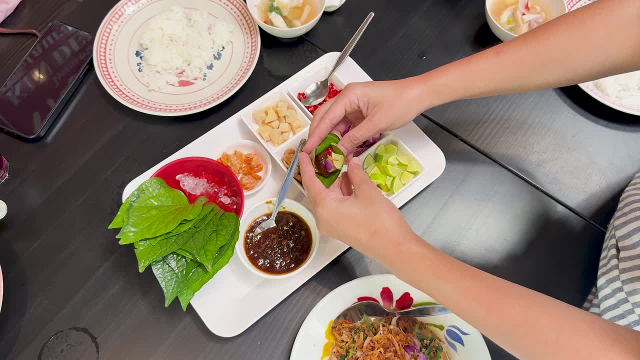 Hands assemble Miang Kham wraps with fresh betel leaves, assorted fillings, and sauce on a modern dining table under bright, even lighting from an overhead perspective