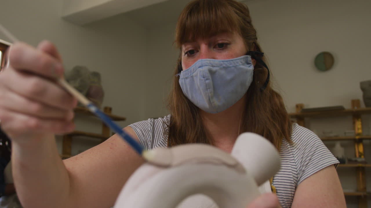 여성 도자기는 얼굴 마스크와 앞옷을 착용하고 도자기 스튜디오에서 도자기에 그림을 그리기 위해 글레이즈 브러쉬를 사용합니다.
