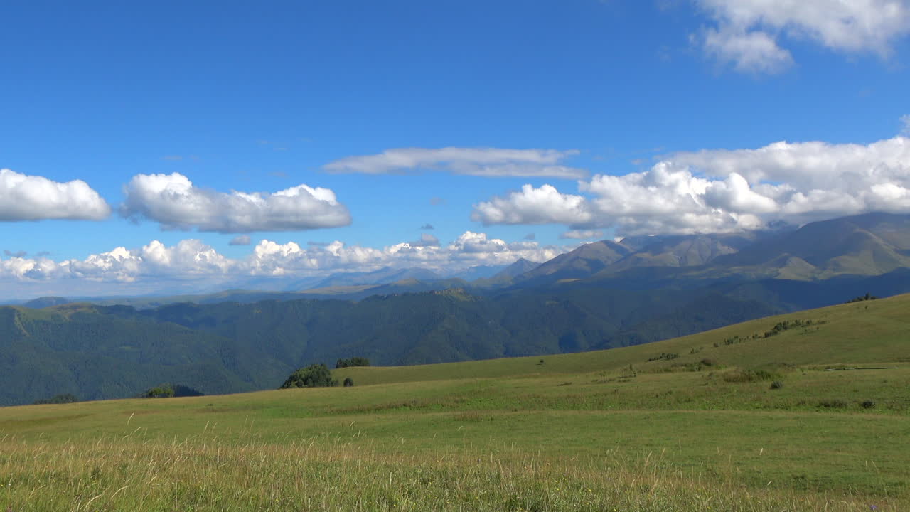 ロシア、コーカサス山脈ドンバイ。遠くの山並みと積乱雲が広がる広大な高山牧草地の、美しい夏の風景映像。