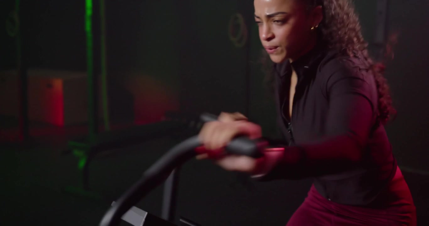 Donna afroamericana con una maglia con cerniera in palestra durante un intervallo, che afferra e spinge le maniglie di una cyclette ad aria, allenandosi.