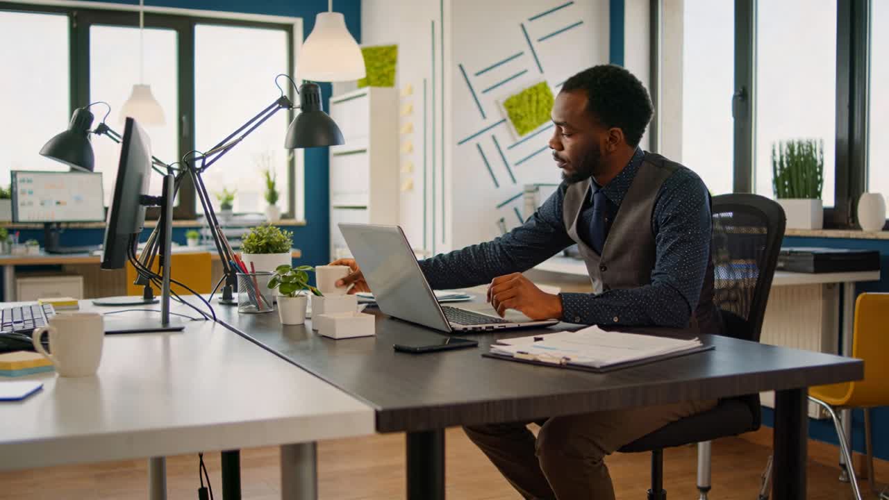 Typing worker in gray vest on laptop at office, adjusting box, sipping mug, resuming, copy space