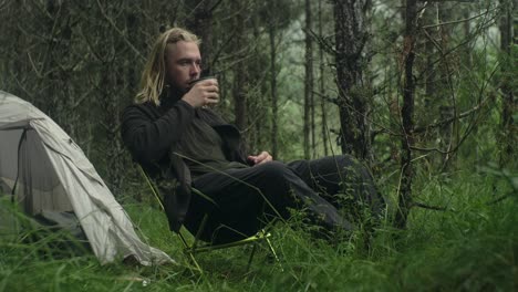 young long haired, bearded, caucasian man solo camping in the woods, sitting drinking coffe