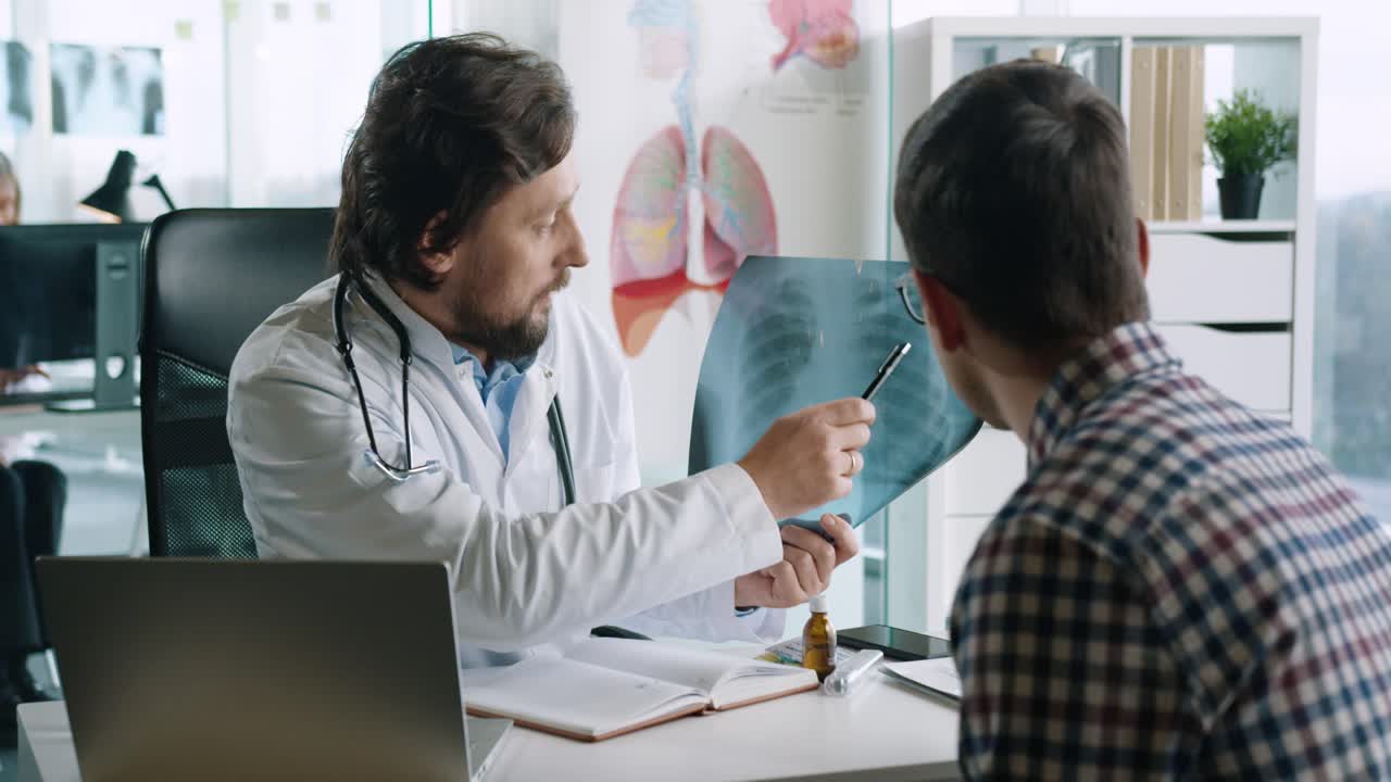vista de primer plano del médico caucásico sentado en el escritorio con una computadora portátil y explicando a un paciente masculino la enfermedad pulmonar por coronavirus en consulta médica