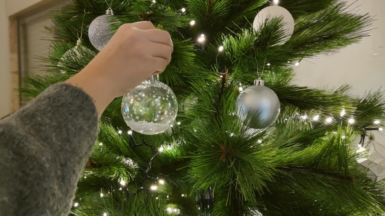 mano de mujer decorando el árbol de navidad dentro de la casa - colgando una bola de navidad clara en el árbol durante la temporada de navidad