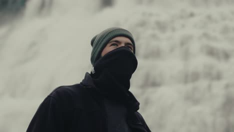 hombre joven guapo con sombrero de invierno y calentador de cuello mirando a la cascada islandesa