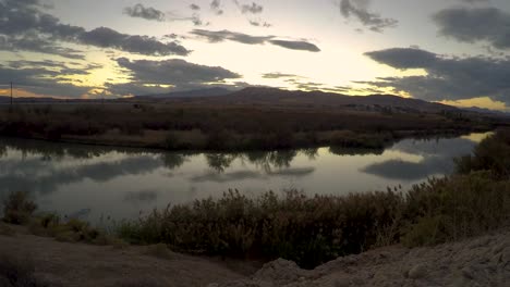 Static-time-lapse-of-a-sunset-cloudscape-reflecting-off-the-smooth-surface-of-a-river-with-mountains-in-the-background