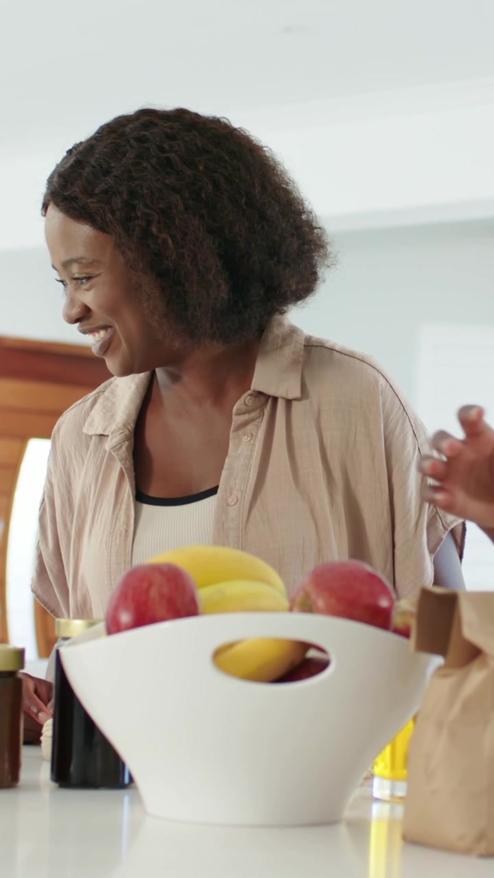 Vídeo vertical: Mulher sorridente com blusa e regata bege na cozinha recebendo ajuda enquanto uma mão coloca uma maçã.