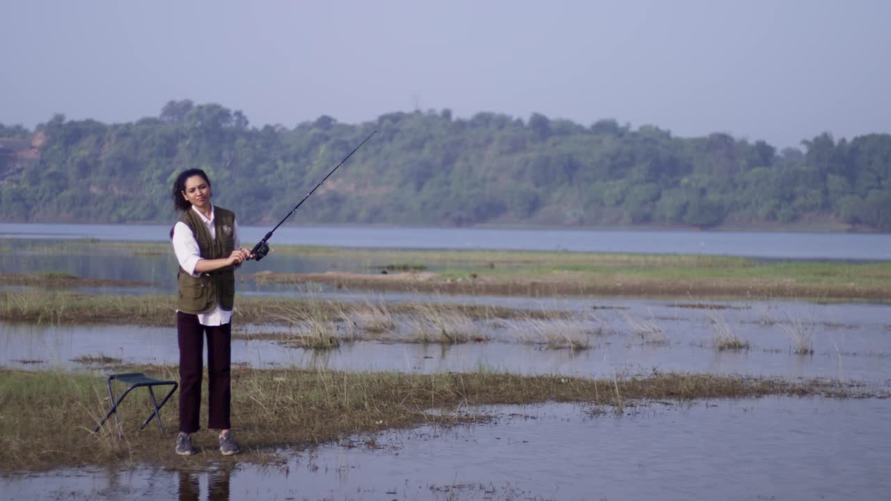 Seorang wanita Asia Selatan tersenyum sambil mengangkat pancing di dekat kursinya di tepi danau yang tenang, menikmati alam pedesaan, air dangkal, dan cahaya siang hari di luar ruangan.