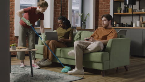 Afroamerican Girl Using Laptop And Boy Playing Video Games Sitting On Sofa While Their Roommate Cleaning The House