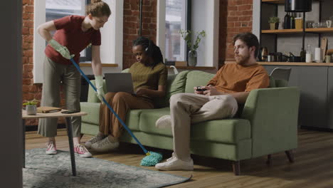 afroamerican girl using laptop and boy playing video games sitting on sofa while their roommate cleaning the house