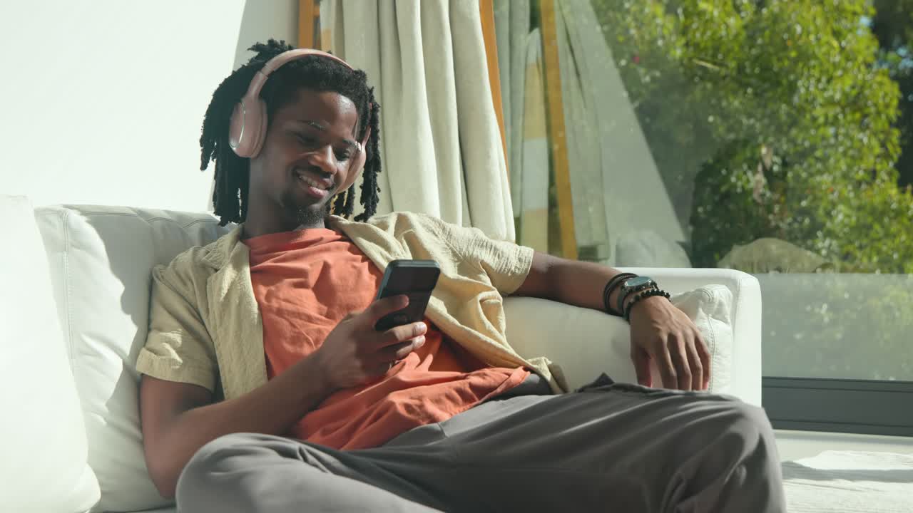Uomo afroamericano adulto con cuffie rosa sdraiato a casa che scorre il telefono reagendo ai contenuti.