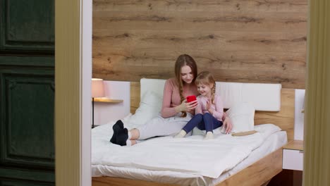 Madre-Acostada-En-La-Cama-Con-Su-Hija-Sosteniendo-Un-Teléfono-Móvil-Haciendo-Compras-En-Línea,-Celebrando-Ganar