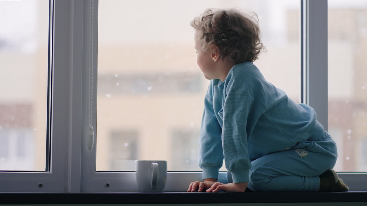 happy and carefree childhood little boy in blue pajamas is sitting on windowsill in apartment in winter day