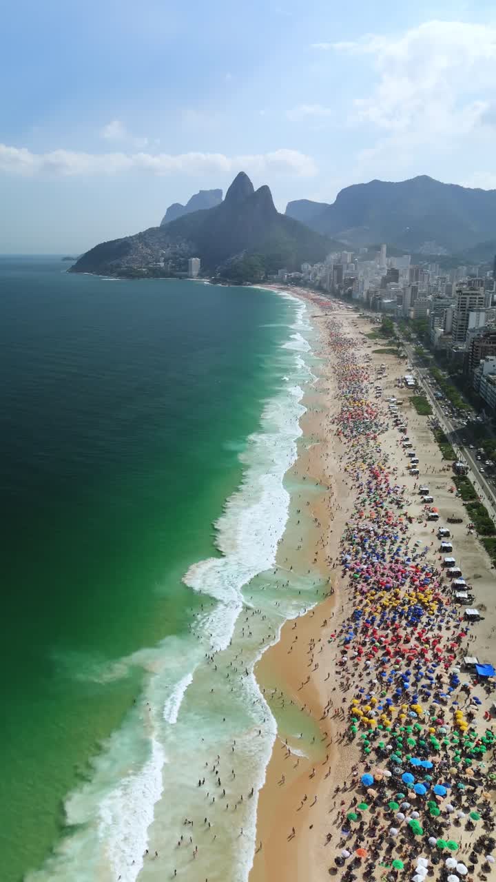 Vertical drone flyover shows Ipanema’s umbrella-packed beach, emerald surf, Dois Irmãos