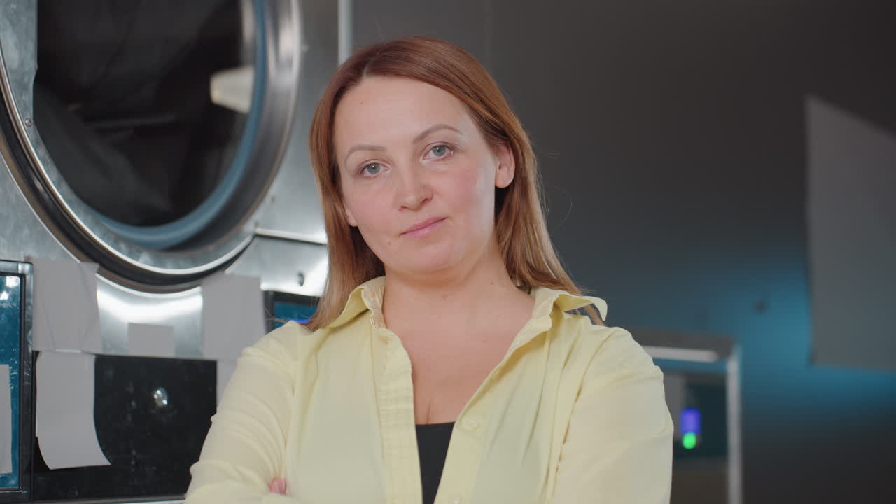 Portrait of stewardess posing near industrial washing machine in laundromat, female worker in yellow shirt looking aside, stainless drum, blue control screens, service backdrop, indoor scene