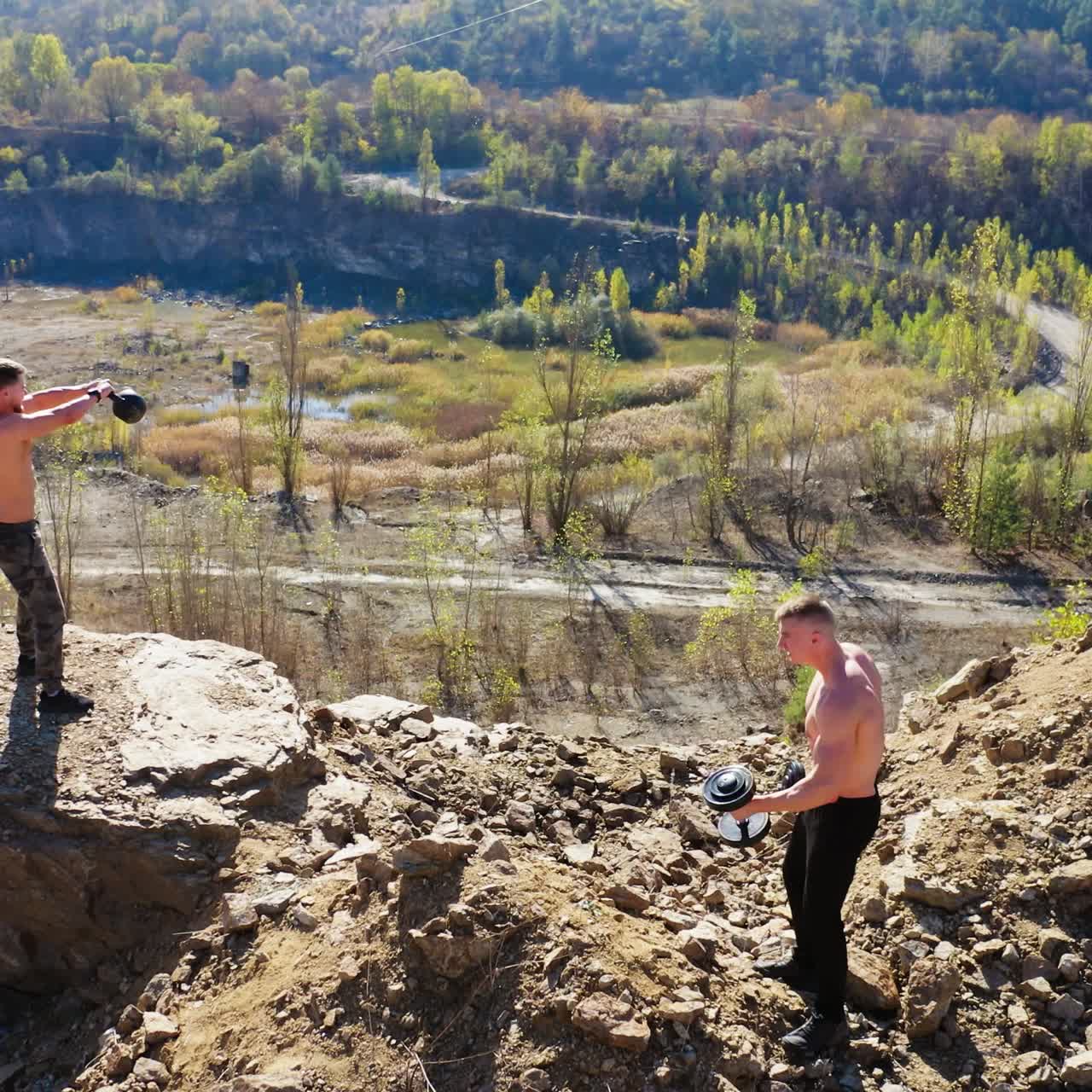 Sportsmen training with sports equipment among nature. Two muscular athletes doing exercises with dumbbells on the top of a hill. Aerial view.
