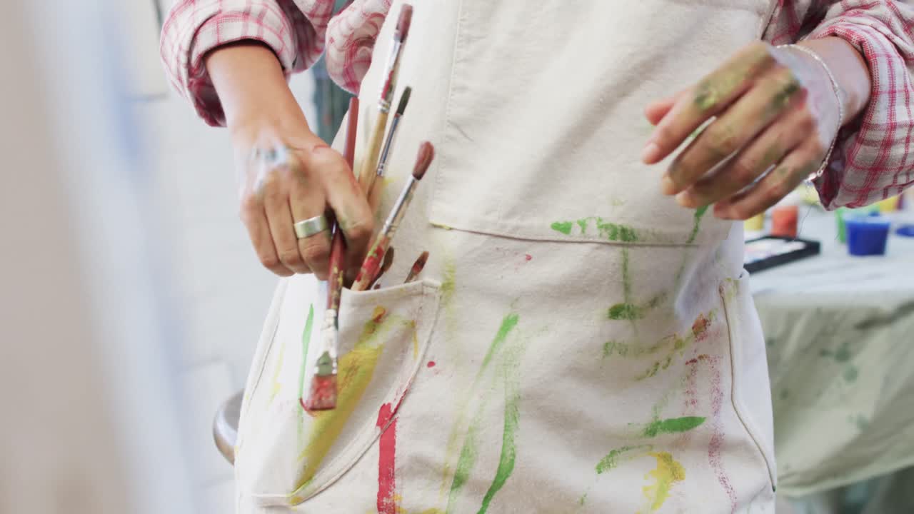 sección media de artista femenina biracial poniendo pinceles de pintura en el bolsillo en el estudio, cámara lenta